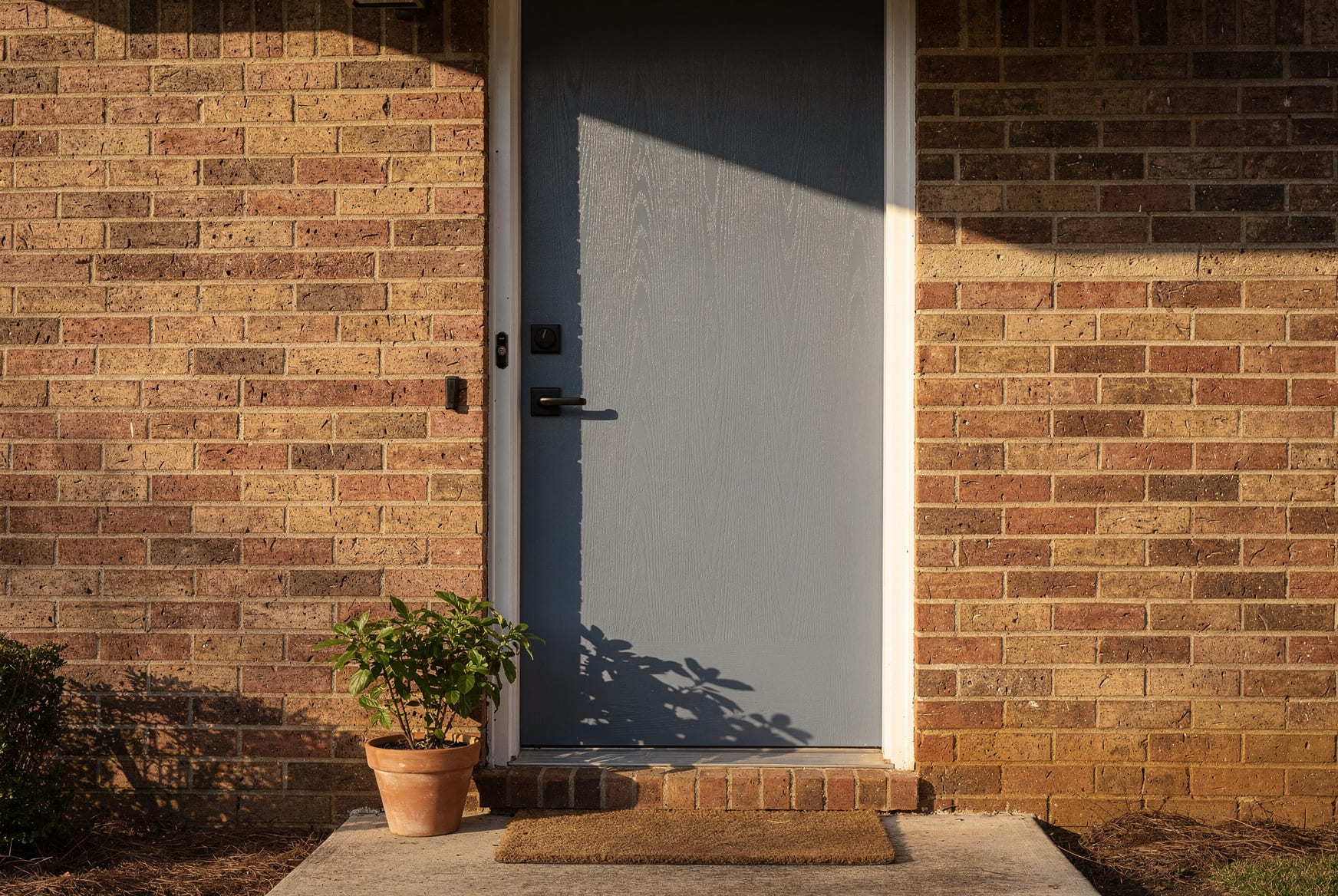 A slate-blue fiberglass entry door on a brick NC home catching warm mid-morning sun on a south-facing wall, the front stoop lit and lived-in, a real light rake across the door face.