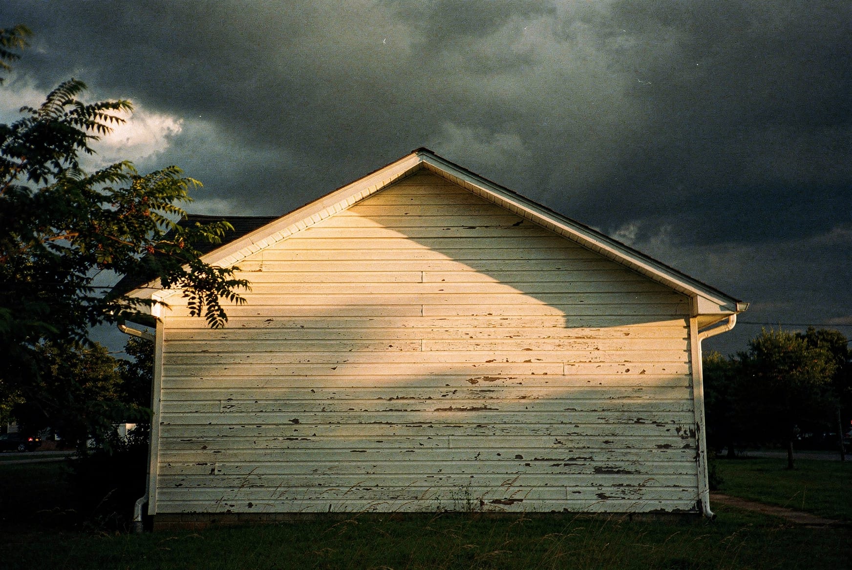 A dark summer thunderstorm rolling in over the sided gable end of a North Carolina home, low warm sun breaking through beneath the storm clouds.