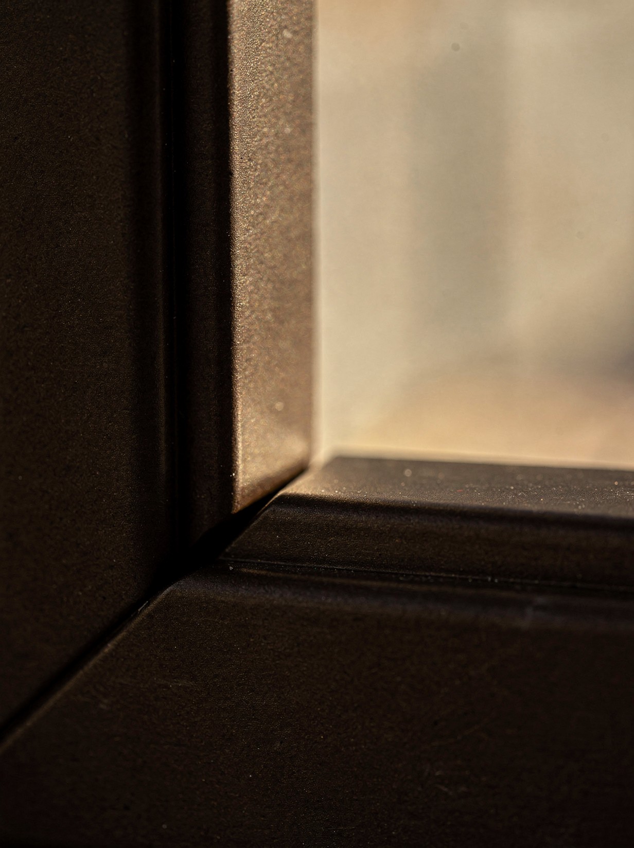 A close macro shot of a dark-bronze composite window frame corner joint, warm late light grazing the matte surface to show its color depth.