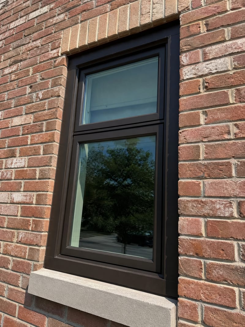A deep-bronze fiberglass window frame in a sun-blasted south-facing brick wall, its frame lines dead straight in hard mid-afternoon light.