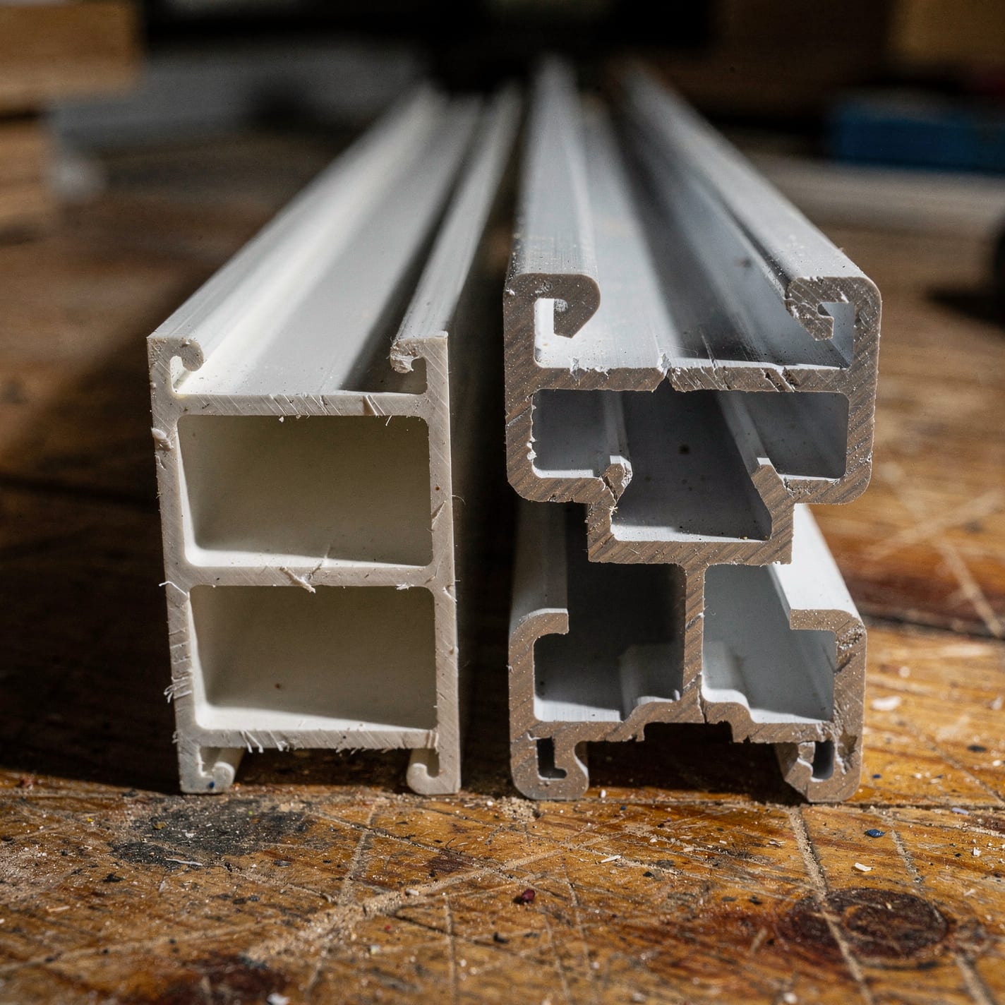 Two cut vinyl window-frame extrusion cross-sections side by side on a workbench — a thin hollow builder-grade profile next to a thicker multi-chambered premium profile.