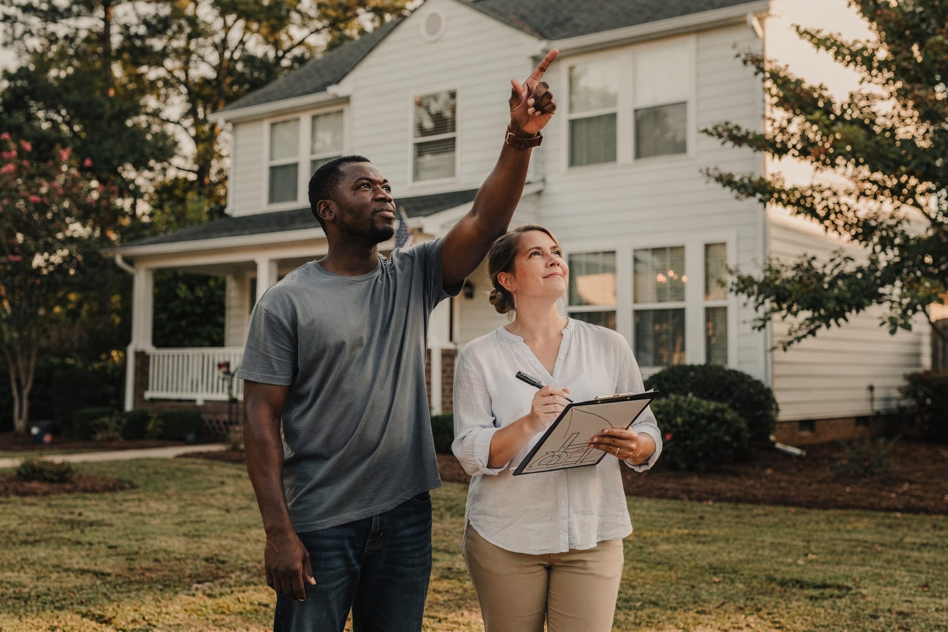 A Black man and a white woman in their forties stand on the front lawn of their two-story North Carolina home during a whole-house walkthrough — he points up at an upper-story window while she marks a clipboard, counting the openings in warm afternoon light.