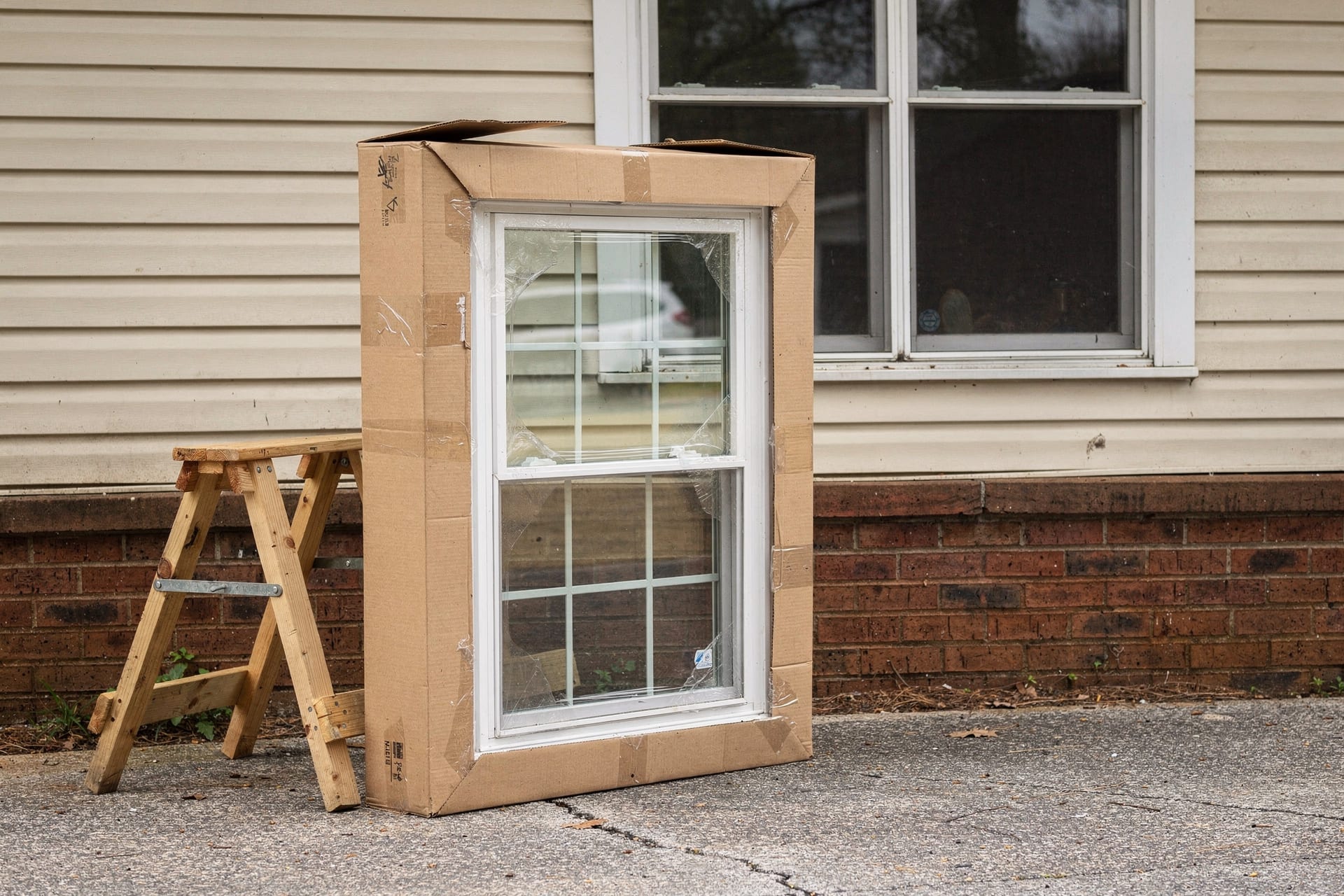 A new replacement window, still in its opened shipping box, leans against a sawhorse on the driveway beside the house — while the old window it will replace sits intact and undisturbed in its opening behind it.