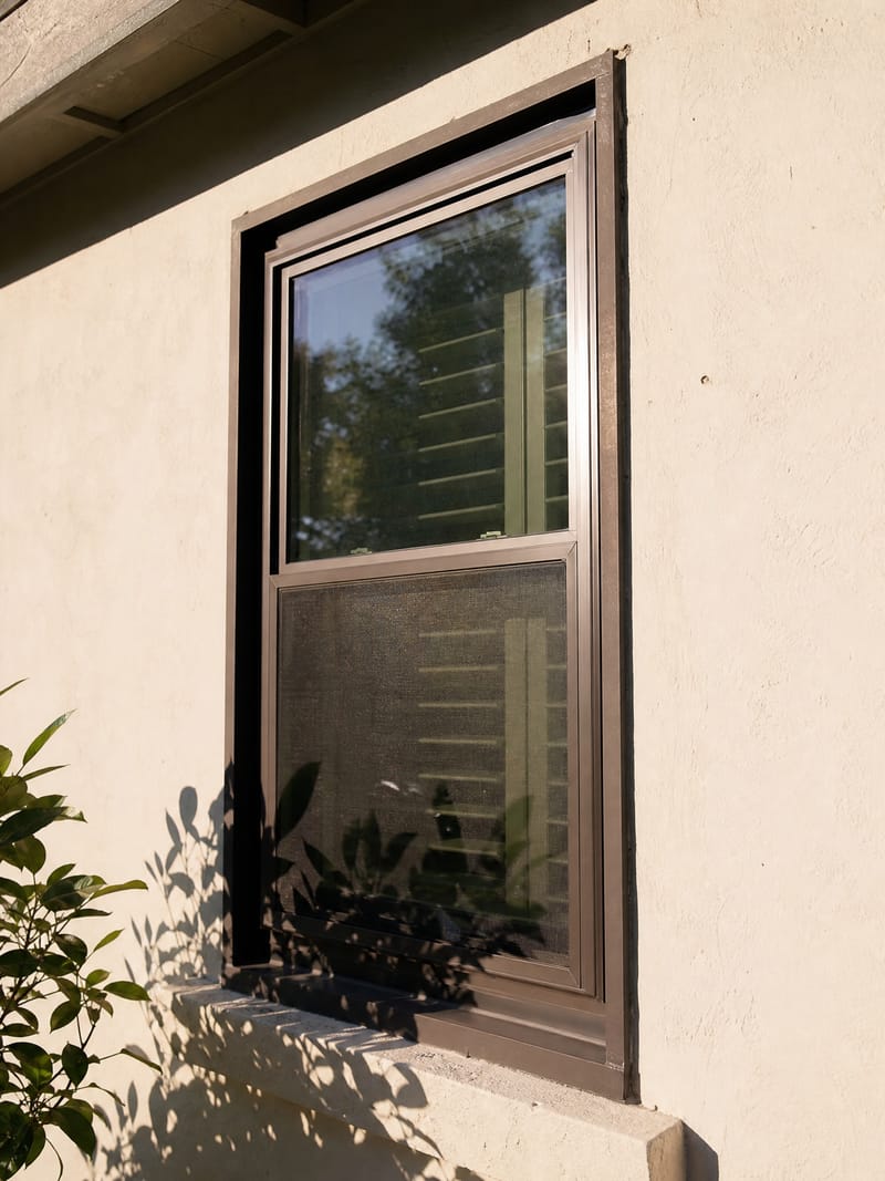 A dark-bronze vinyl window sash in a bright, west-facing wall, baking in hard late-afternoon summer sun.