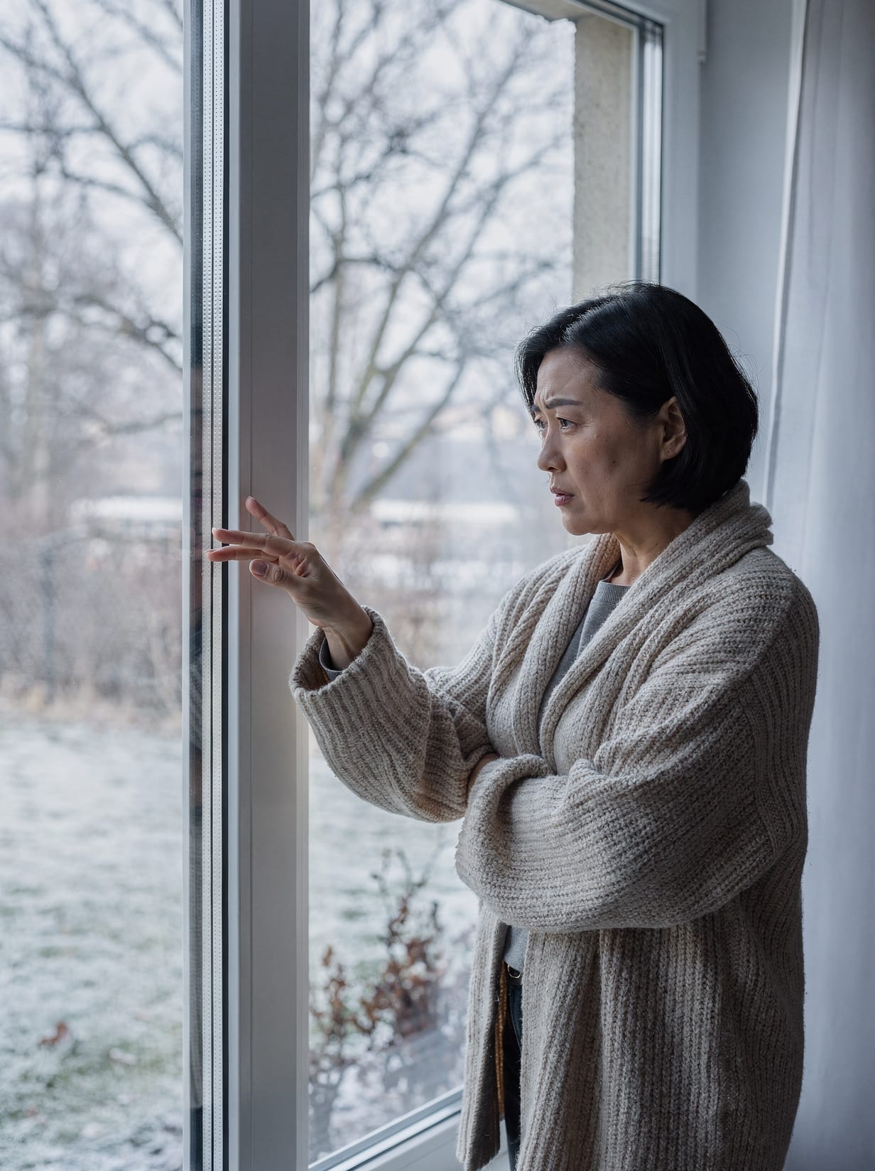 A homeowner wrapped in a cardigan holds a hand up to a clean, modern window on a frosty winter day, frowning at the cold air pouring off the glass.